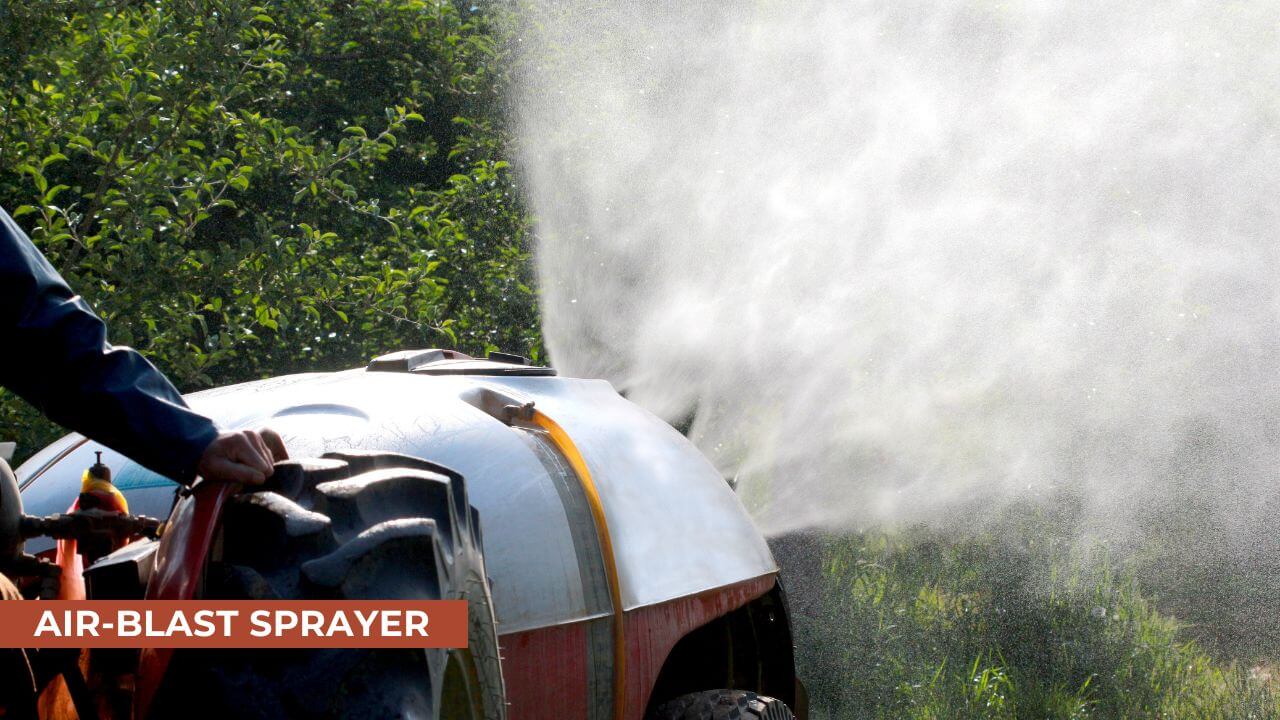 A tractor-powered air-blast sprayer emitting a fine mist of water or chemicals in an orchard, surrounded by green trees and foliage on a sunny day