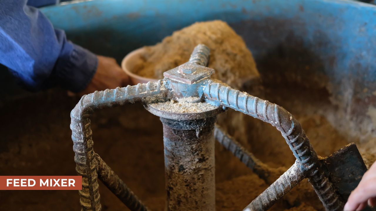 Close-up of a feed mixer machine mixing animal feed, with a person adding material into the mixer and rotating augers visible in the image