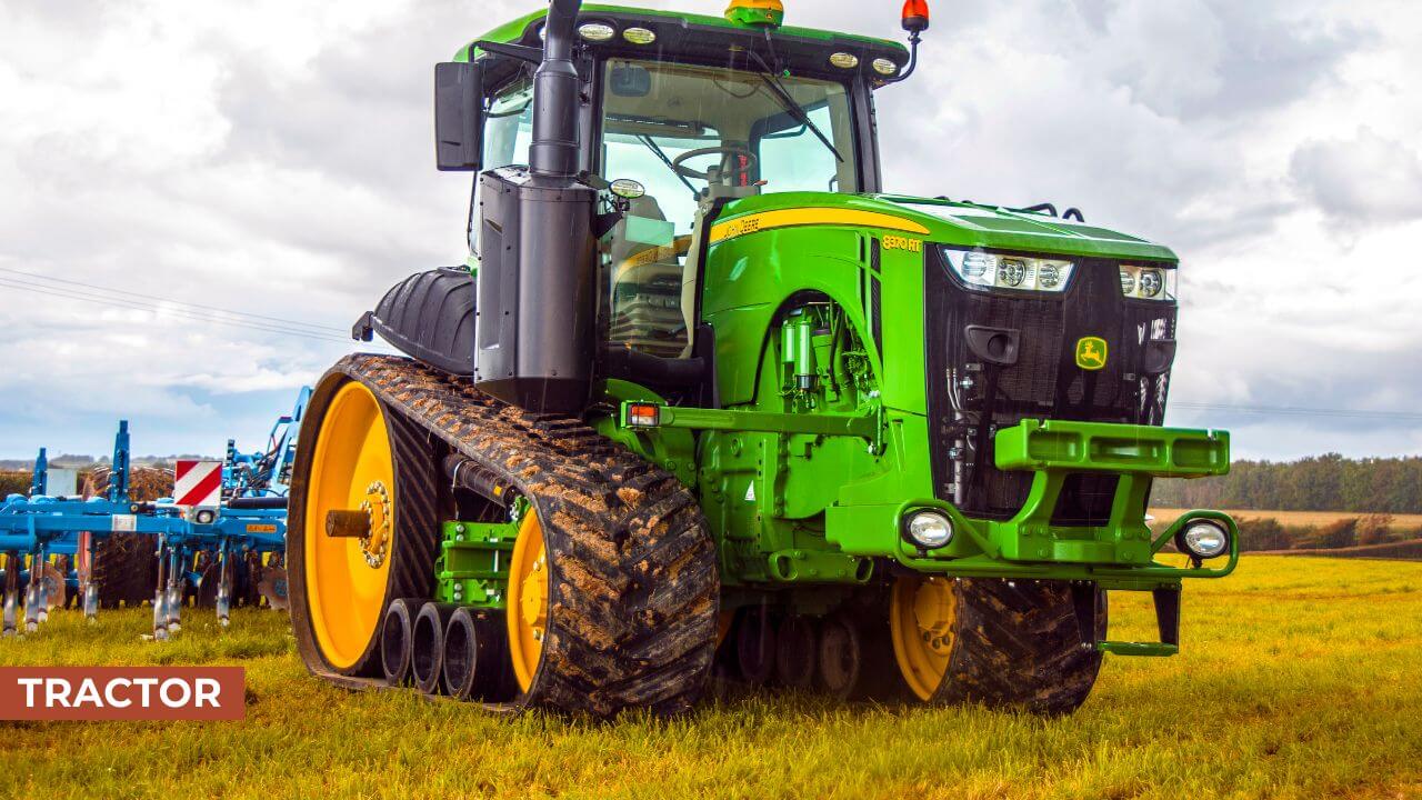 A green and yellow John Deere tractor with wide tracks, standing in a field, ready for agricultural work with a plow attachment in the background